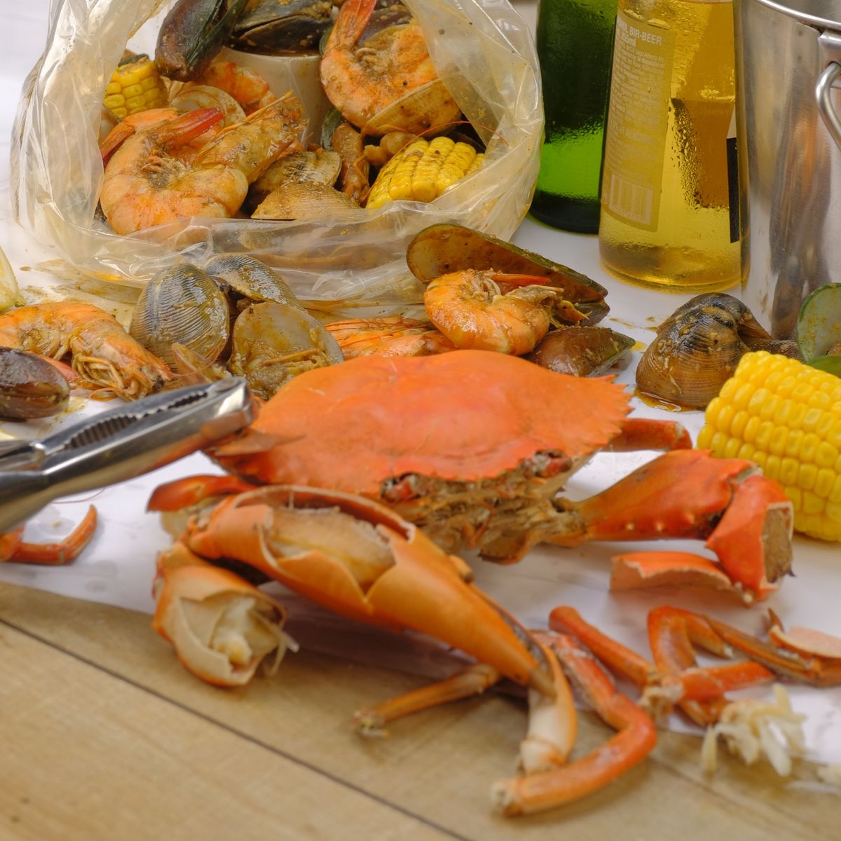 A whole boiled crab with cracked claws beside an opened bag of shrimp, mussels and clams, cold beer behind
