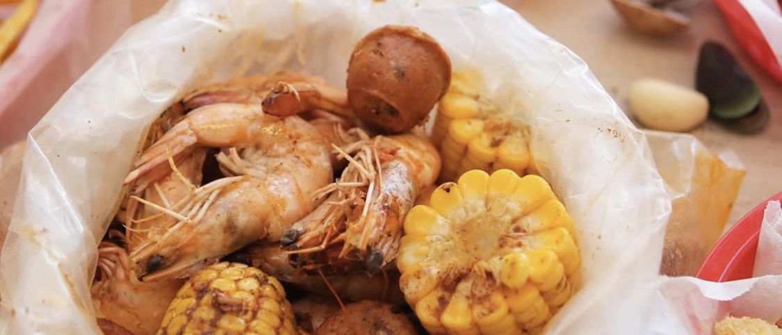 A bag of shrimp, corn and sausage on the table with baskets of lumpia and sides around it