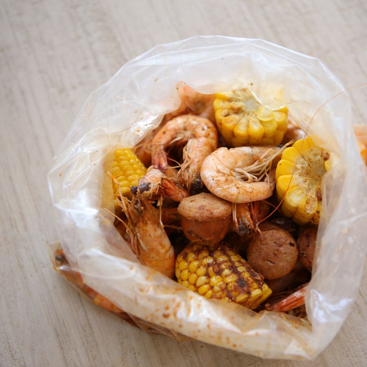 An opened bag of head-on shrimp, corn and sausage dusted in Cajun spice on a pale wooden table